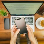 Image of hand holding phone with laptop in the background.